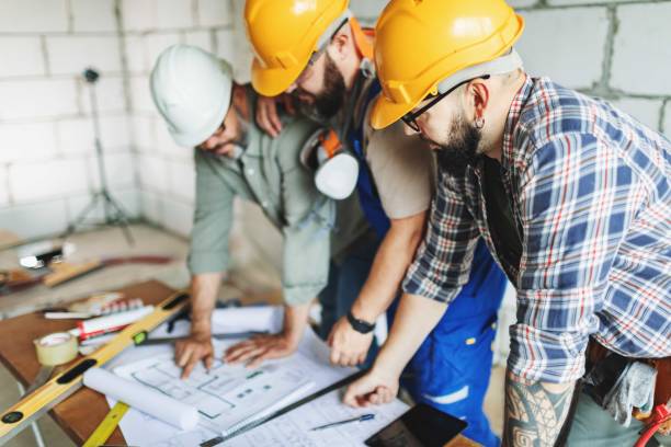 Three manual construction workers in hard hats discussing blueprints at a construction site using a laptop during complete home remodeling in Hillsborough, CA.